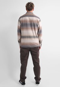 Camicia a maniche lunghe con bottoni di colore beige, con righe orizzontali marroni, grigie e crema. Abbinata a pantaloni marroni scuri e sneakers nere.