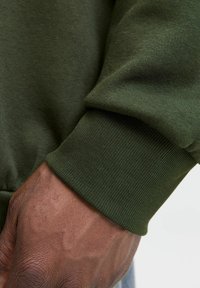 Nærbillede af grøn sweatshirtstof med ribbede manchetter og en blød tekstur med en subtil glans, der fremhæver en tætsiddende pasform om håndleddet.