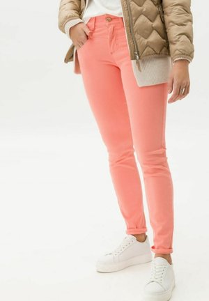 Persoon draagt koraalkleurige slim-fit jeans, witte sneakers, een beige doorgestikt jack en een wit topje, met één hand in de zak.