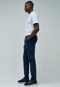 Hombre de pie de lado vistiendo una camiseta blanca, pantalones deportivos azul marino y zapatillas azul marino frente a un fondo gris claro liso.