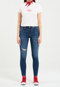 Camiseta blanca corta con gráfico rojo, jeans ajustados azules con una rodilla deshilachada, y zapatillas altas rojas. Fondo liso.