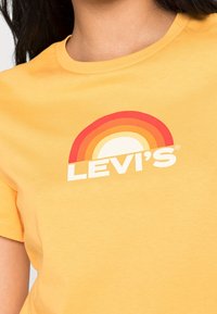 Žluté bavlněné tričko s potiskem duhy v červené, oranžové a bílé barvě, s nápisem "LEVI'S" vytištěným pod grafikou tučnými bílými písmeny.