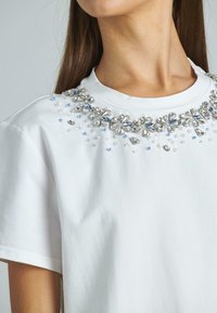Camiseta de algodón blanca con un cuello decorativo que presenta adornos claros y azul claro, incluyendo formas de flores y detalles similares a joyas.