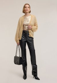 Cárdigan de punto beige con tejido abierto, top metálico color crema, pantalones de cuero ecológico negros de cintura alta y un bolso negro con correa de cadena.