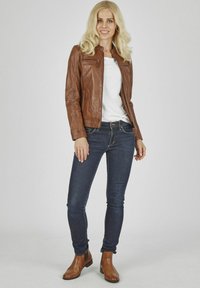 Braune Lederjacke mit figurbetontem Design, Reißverschlusstasche, über einem weißen Hemd getragen. Kombiniert mit dunkelblauen Jeans und braunen Knöchelstiefeln.