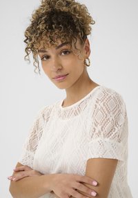 Haut en dentelle blanche à manches courtes, doté d'un motif en losange et d'un col rond. Texture douce avec des détails transparents le long des manches.