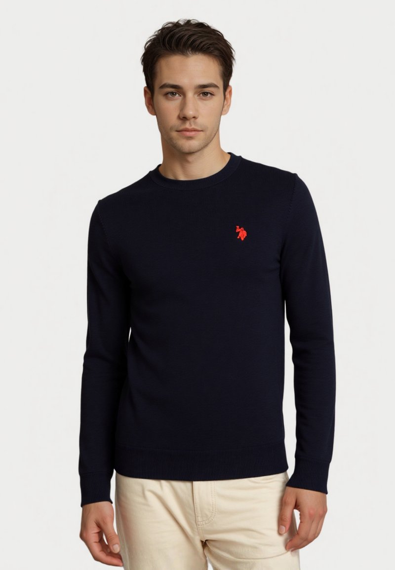 Giovane uomo che indossa un maglione a maniche lunghe blu navy con logo rosso sul petto e pantaloni beige, in piedi davanti a uno sfondo bianco semplice.