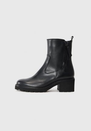 Bottine en cuir noir avec fermeture éclair latérale, talon épais et languette à l’arrière sur fond gris clair uni.