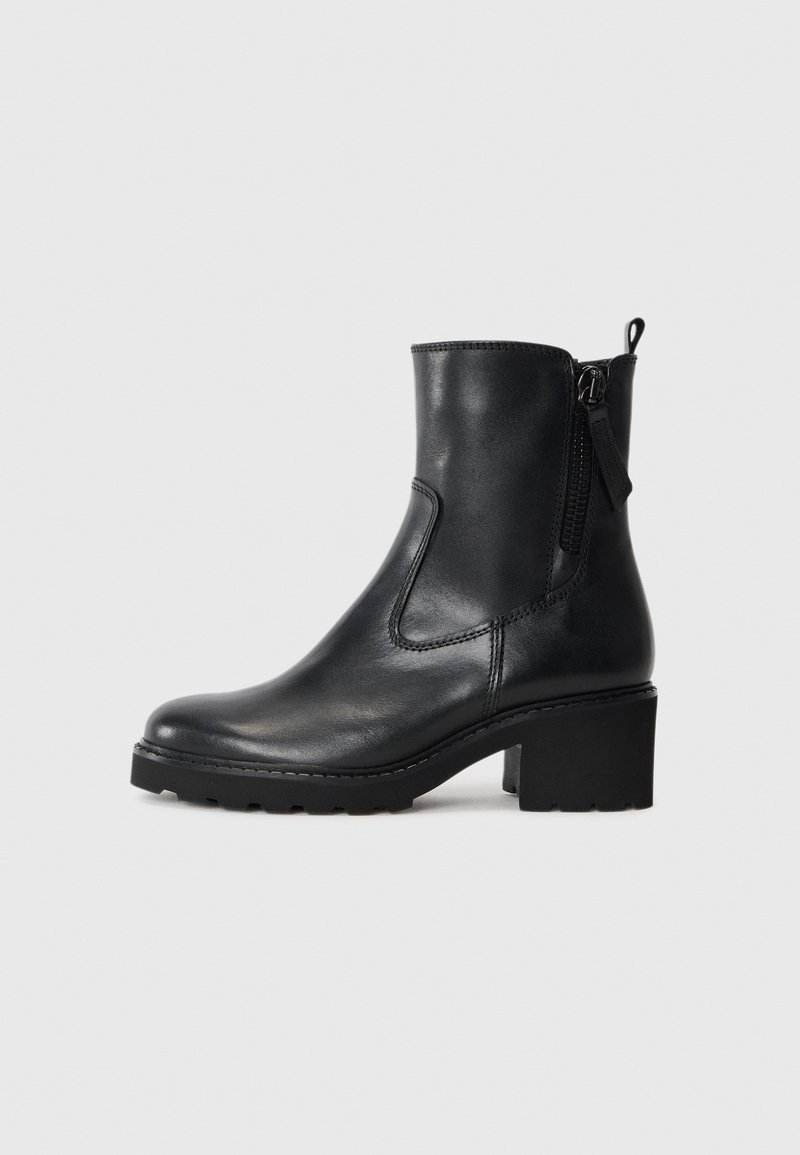 Bottine en cuir noir avec fermeture éclair latérale, talon épais et languette à l’arrière sur fond gris clair uni.
