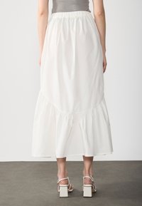Jupe maxi blanche à volants avec une taille froncée et une silhouette fluide, fabriquée en tissu léger. Portée avec des sandales à talons épais.