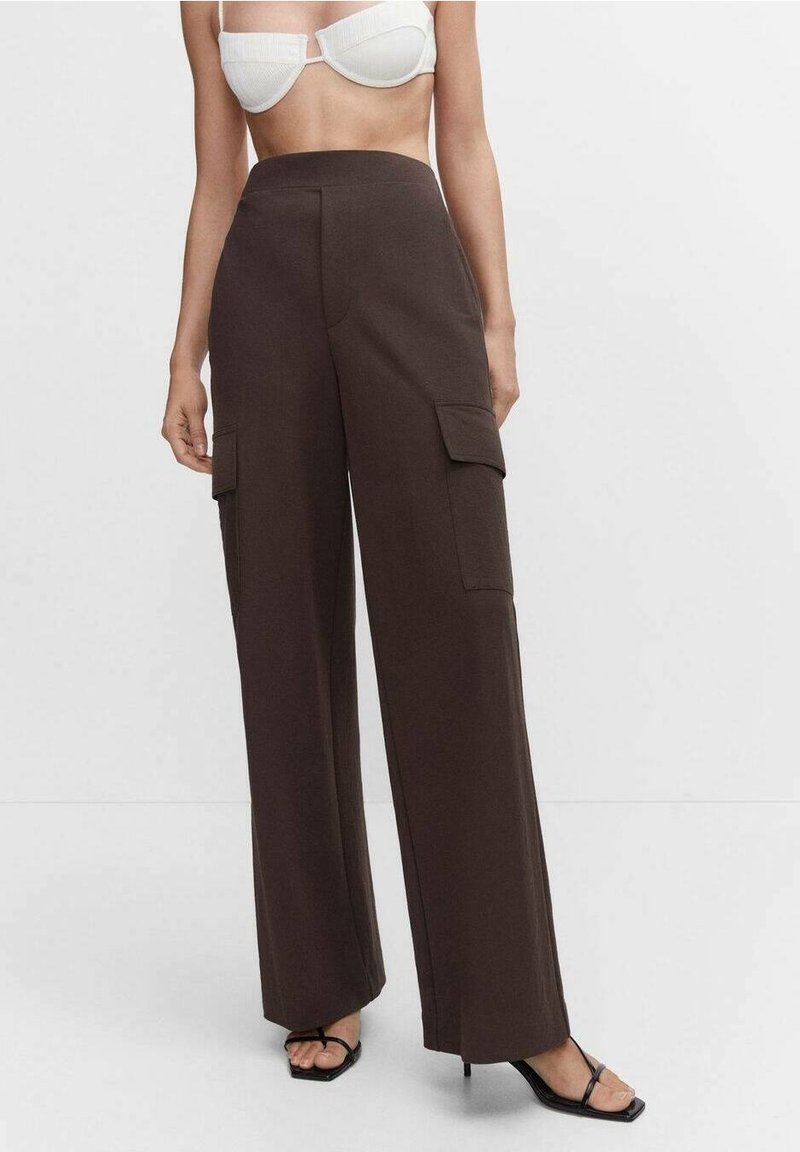 Femme portant un pantalon large taille haute marron foncé avec des poches cargo latérales, des sandales à talons noires à brides, et un haut court blanc visible.