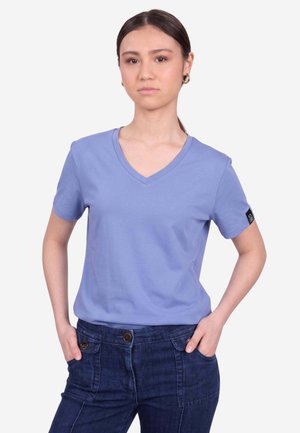 Giovane donna con capelli scuri raccolti in una coda di cavallo, che indossa orecchini a cerchio dorati, una maglietta viola chiaro con scollo a V e jeans blu scuro con le mani nelle tasche.