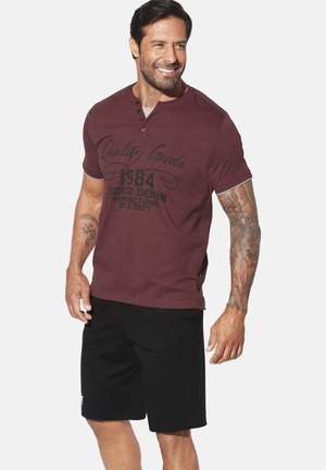 Burgundy short-sleeve polo shirt with button placket, featuring printed graphics in black. Paired with black shorts in a soft fabric.