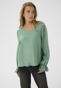 Top verde chiaro a maniche lunghe con scollatura a V e polsini arricciati. Realizzato in tessuto morbido, presenta una vestibilità comoda e una consistenza liscia.
