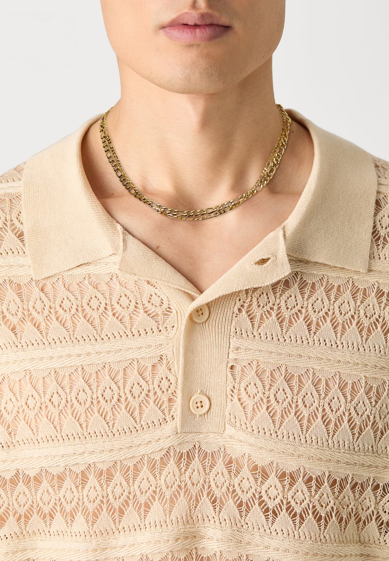 Camisa polo de punto beige con un patrón parecido al encaje, cuello corto, tres botones y un collar de cadena dorada.