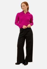 Fuksjowy sweter robiony na drutach z dekoltem w kształcie V i kołnierzem, w zestawieniu z szerokimi czarnymi spodniami z przednimi kieszeniami, noszonymi z czarnymi butami.