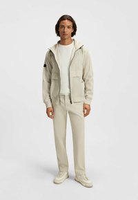 Chaqueta de cremallera beige claro con capucha, combinada con un suéter blanco texturizado, pantalones a juego y zapatillas beige. Diseño informal, con acentos mínimos.