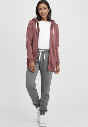 Burgunderfarget zip-up hettegenser med hette og hvite snorer, brukt over en hvit t-skjorte; kombinert med grå joggebukser og hvite joggesko.
