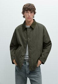 Olijfgroene canvas jas met een gestructureerde kraag, drukknopen sluiting en zijzakken, gecombineerd met een grijs T-shirt en lichtblauwe spijkerbroek.