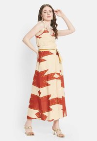 Robe maxi beige avec des motifs géométriques rouges, dotée d'une taille nouée, d'un décolleté en V et de bretelles volantes. Le mannequin porte des sandales métalliques.