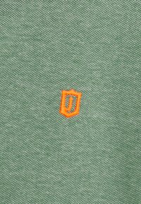 Grønt tekstil med en strukturert overflate og et lite oransje skjoldformet logo med en stilisert "J" i sentrum.