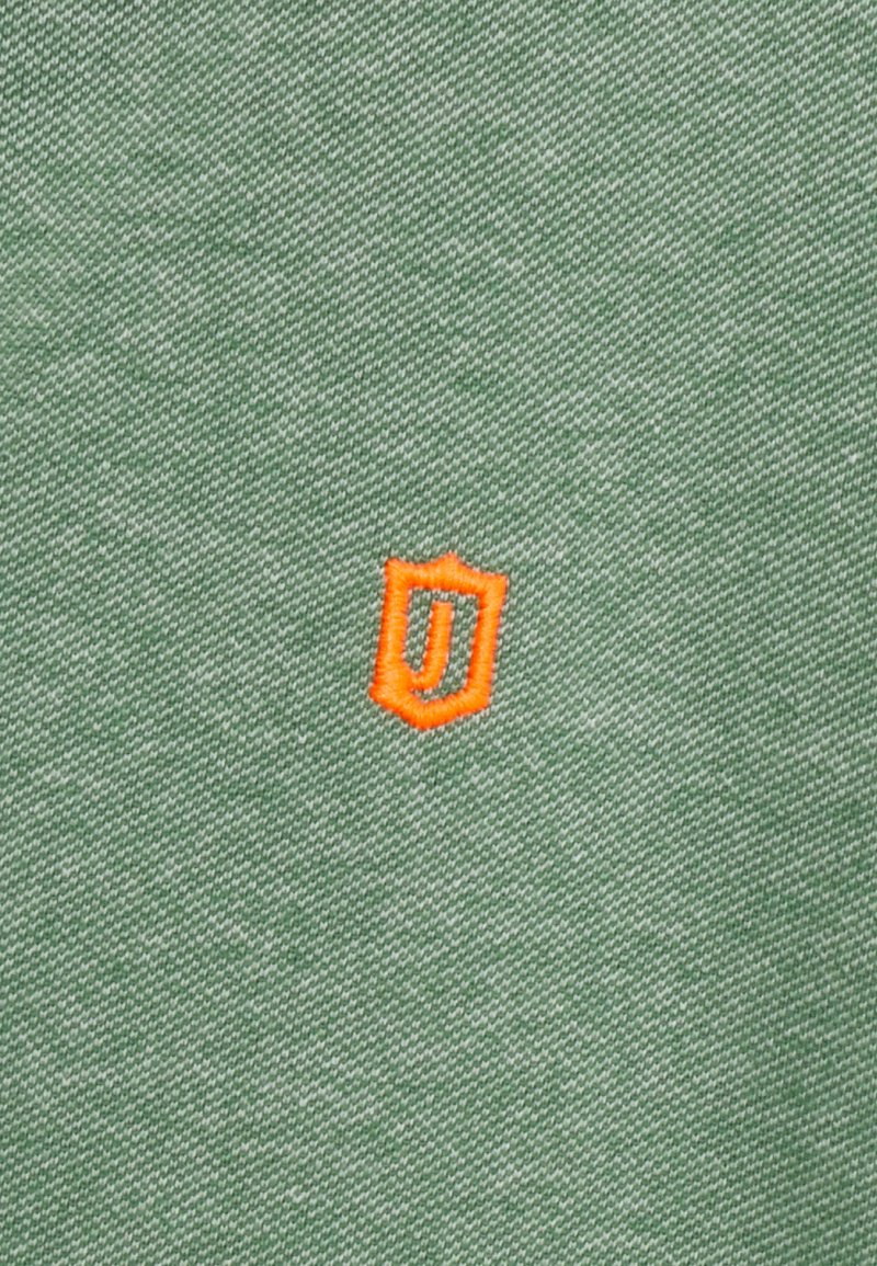 Grüner Stoff mit einer strukturierten Oberfläche und einem kleinen, orangefarbenen, schildförmigen Logo, das in der Mitte ein stilisiertes "J" zeigt.