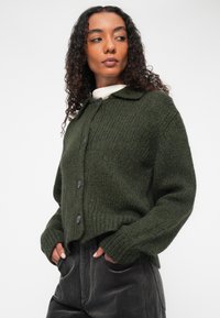 Cardigan en laine vert foncé avec un col, des poignets côtelés et un boutonnage à l'avant. Motif tricoté texturé ; design court.