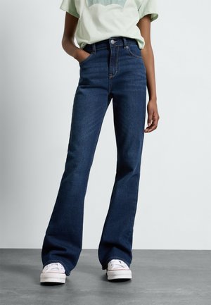 Personne portant un jean flare taille haute bleu foncé, un t-shirt vert clair et des baskets blanches, debout avec une main dans la poche sur un sol gris.
