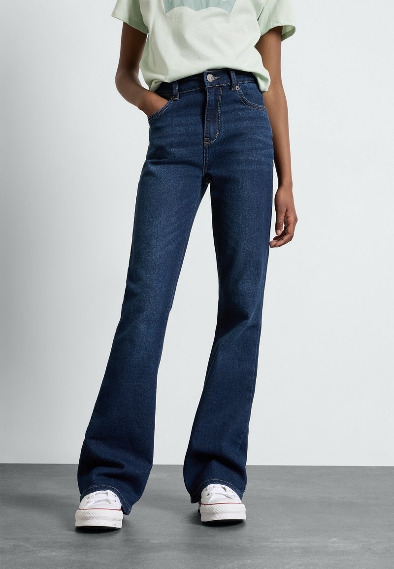 Personne portant un jean flare taille haute bleu foncé, un t-shirt vert clair et des baskets blanches, debout avec une main dans la poche sur un sol gris.