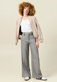 Beige bomberjack over een wit T-shirt, gecombineerd met hooggerende, wijde grijze broek en witte sneakers met zwarte strepen.