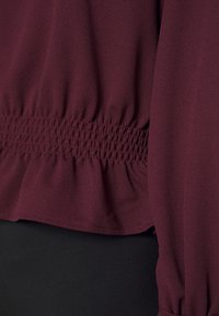 Bordeauxfarbene Bluse mit smocktem Taillenbund, langen Ärmeln und weichem, leichtem Stoff. Peplum-Saum mit gerafftem Rand.