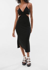 Vestido negro de punto con un lazo en la parte delantera y lados recortados, que cuenta con una abertura alta en el muslo. Se combina con sandalias de plataforma blancas y gruesas.