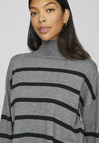 Pilkas megztas turtleneck svarkas su juodomis horizontaliomis juostelėmis, pasižymintis laisvu siluetu ir raižyta tekstūra ties kaklu.