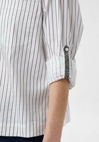 White shirt with black vertical stripes, featuring a front pocket and rolled sleeves adorned with a leopard print accent on the cuff.