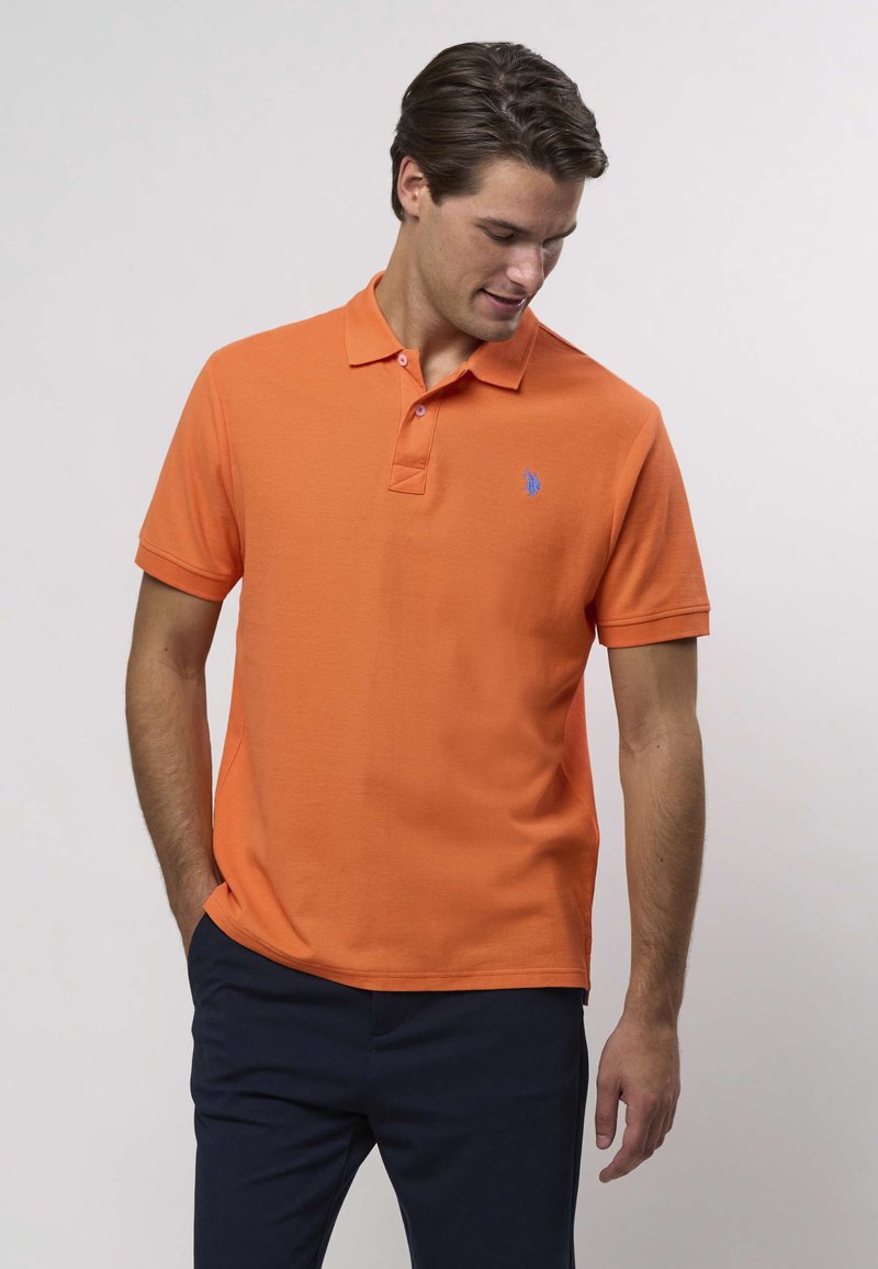 Oranžist polo-särk, mis on valmistatud tekstureeritud kangast, klassikalise krae, kolme nupuga esiosaga ja väikese sinise logoga rinnal.
