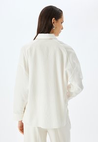 Camisa blanca de manga larga con cuello, de rayas en relieve. Presenta un corte relajado y una textura suave, con patrón de rayas visible y puños enrollados.