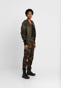 Olijfgroene bomberjack, zwart knopenhemd, camouflagebroeken en zwarte veterschoenen. Gladde stof, losse pasvorm en gemêleerd design.