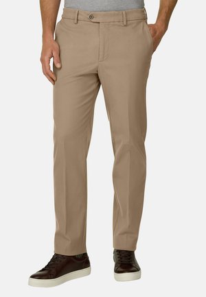 Mand iført beige bukser med lige ben og mørkebrune sneakers med hvide såler, hænder delvist i lommerne, står mod en ensfarvet baggrund.