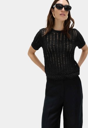 Kvinde iført sort åbent strikket kortærmet top, sorte bukser, store guldfarvede øreringe og sorte solbriller, stående med hænderne på hoften.