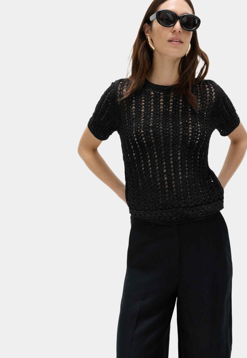 Frau trägt schwarzen kurzärmligen, offen gestrickten Oberteil, schwarze Hose, große goldene Ohrringe und schwarze Sonnenbrille, steht mit Händen in der Hüfte.