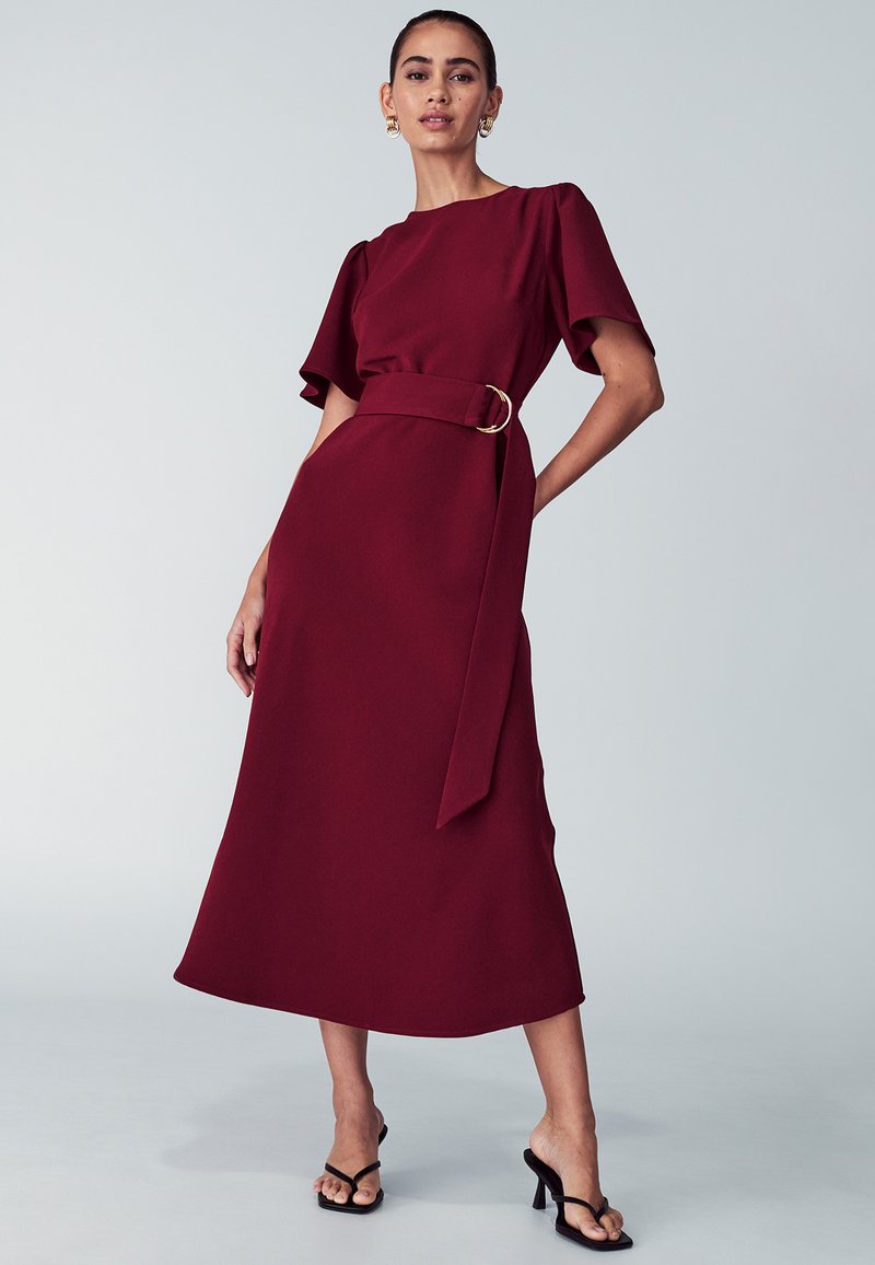 Burgundijas midi kleita ar īsām pūkām, apaļu kakla izgriezumu un jostu viduklī ar zelta toņa apaļu sprādzi. Gluda auduma tekstūra.