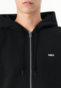 Black zip-up hoodie made of soft fabric, featuring a front zipper, drawstring, and white "OBEY" logo on the left chest area.