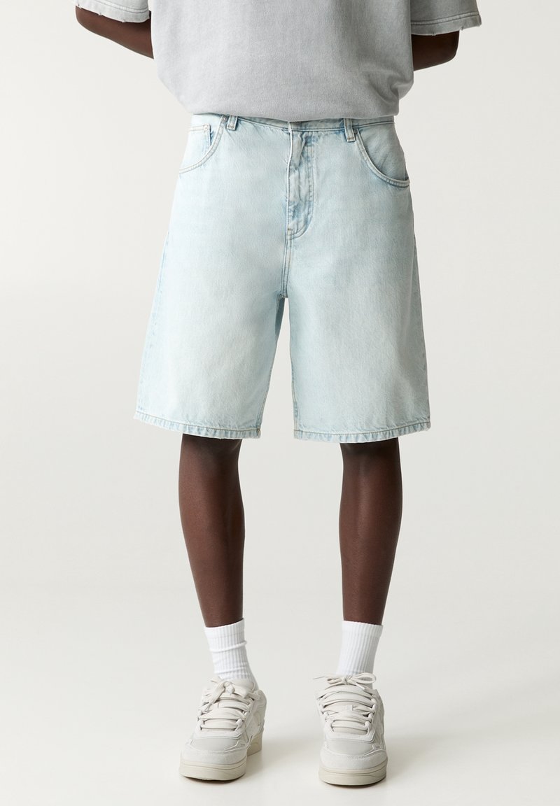 Personne portant un short en denim bleu clair, des chaussettes blanches à mi-mollet et des baskets blanches, se tenant devant un fond uni.