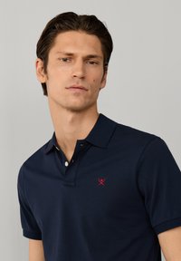 Ung mand med kort mørkt hår iført en marineblå polo med en lille rød broderet logo, neutral lysegrå baggrund.