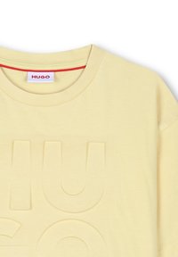 Camiseta de algodón amarilla con cuello redondo, que presenta un logotipo en relieve de "HUGO". Con un acabado en el cuello interior de color rojo. Textura suave.