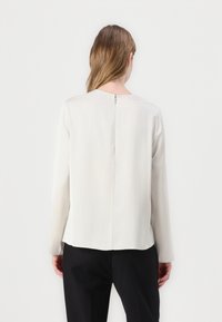 Blouse gris clair à manches longues avec un col rond, fermeture éclair dans le dos et texture de tissu lisse, portée avec un pantalon noir.