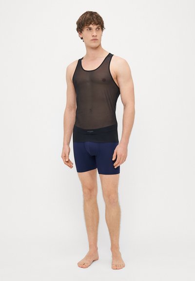 Fekete áttetsző ujjatlan felső széles pántokkal és testhezálló derékrésszel, sötét tengerészkék boxeralsóval párosítva. A modell mezítláb áll egy fehér háttér előtt.