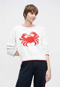 Λευκό πλεκτό πουλόβερ με σχέδιο κόκκινου καβουριού. Ριμπ λαιμόκοψη και τελείωμα με κόκκινες λεπτομέρειες. Χαλαρή εφαρμογή, μακριά μανίκια και απαλό υφή.