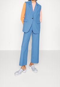 Ljust blå skräddarsydd väst med två fickor, ihop med matchande vida byxor. Bärs över en orange långärmad topp och vita slip-on träskor.