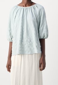 Haut bleu clair à manches bouffantes en tissu texturé, avec un col rond et une coupe décontractée, associé à une jupe blanche fluide.